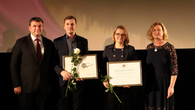 Četri cilvēki, divi no viņiem tur Atzinības rakstus. Divas sievietes, divi vīrieši. Tumšs fons
