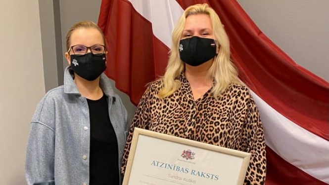 Valsts karogs fonā, LATAK vadītāja un sabiedrisko attiecību speciāliste ar Atzinības rakstu rokās Sandra Kušķe
