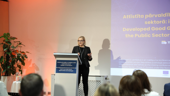 LATAK direktore, sieviete gaišiem matiem, stāv uz skatuves un stāsta par savu tēmu, fonā prezentācijas slaids - zils ekrāns ar dzelteniem burtiem