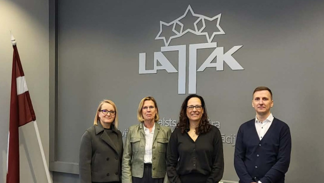 Četri cilvēki, trīs sievietes un viens vīrietis stāv uz LATAK logotipa fona, kreisajā pusē Latvijas valsts karogs