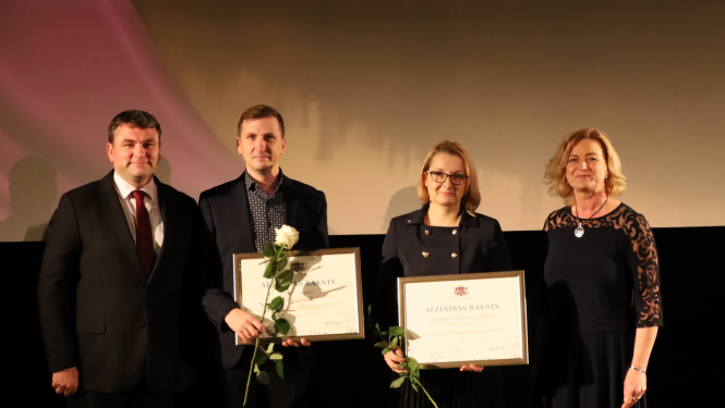 Četri cilvēki, divi no viņiem tur Atzinības rakstus. Divas sievietes, divi vīrieši. Tumšs fons