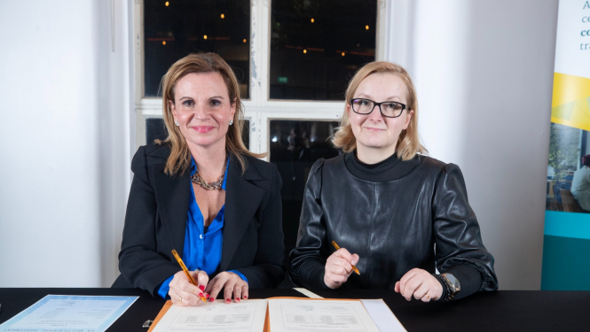 Parakstīšanas brīdis, kurā piedalās LATAK direktore un EA pārstāve. Uz galda ir mape ar dokumentiem un abas pārstāves veic parakstīšanu.