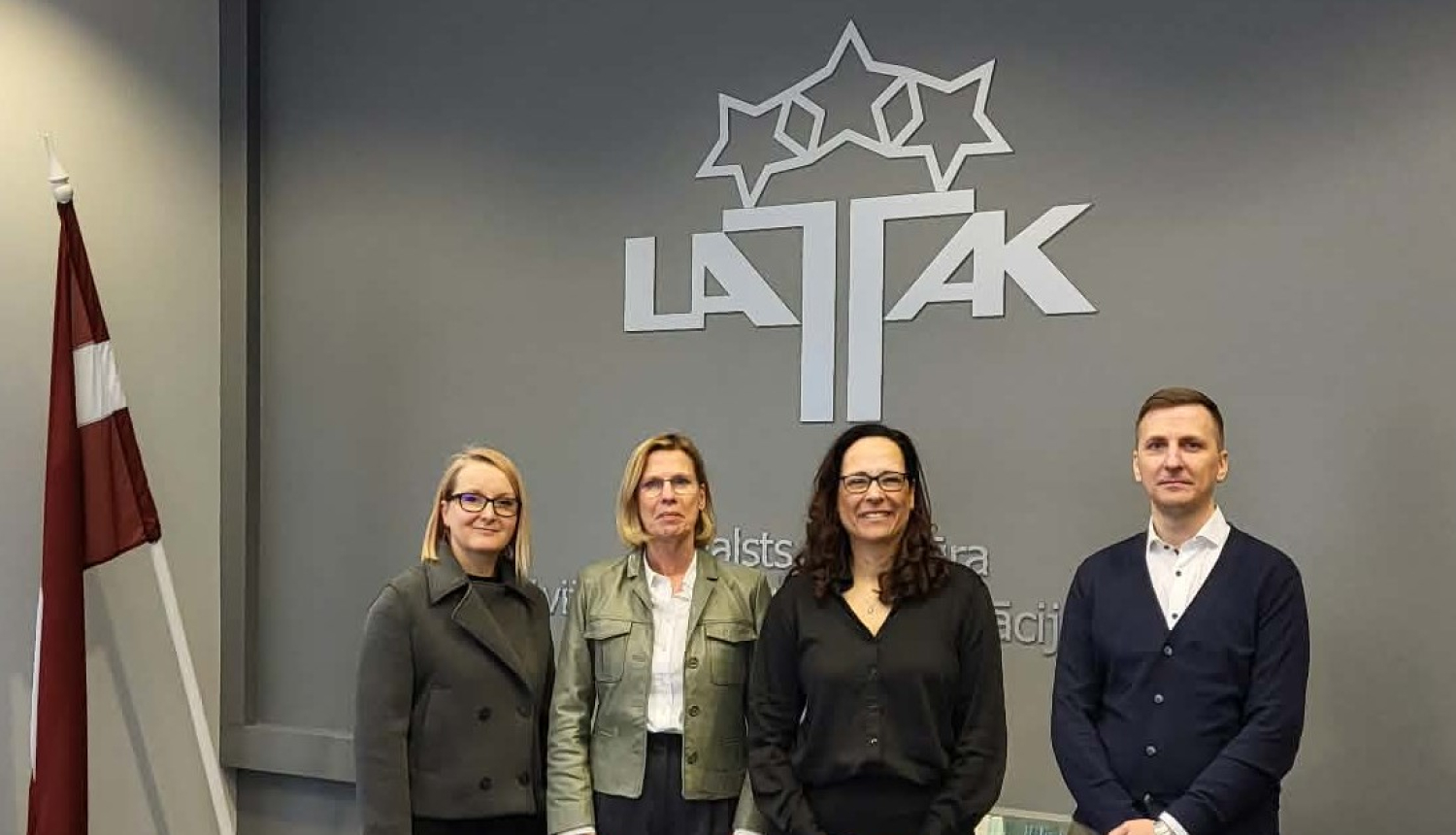 Četri cilvēki, trīs sievietes un viens vīrietis stāv uz LATAK logotipa fona, kreisajā pusē Latvijas valsts karogs