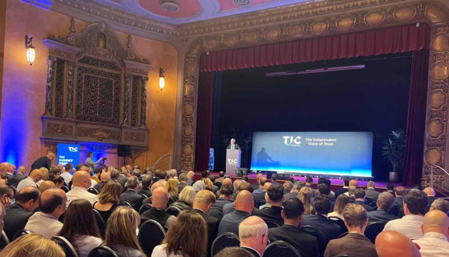 TIC samita skats, zāle ar cilvēkiem, skatuve ar ekrānu un vienu runātāju