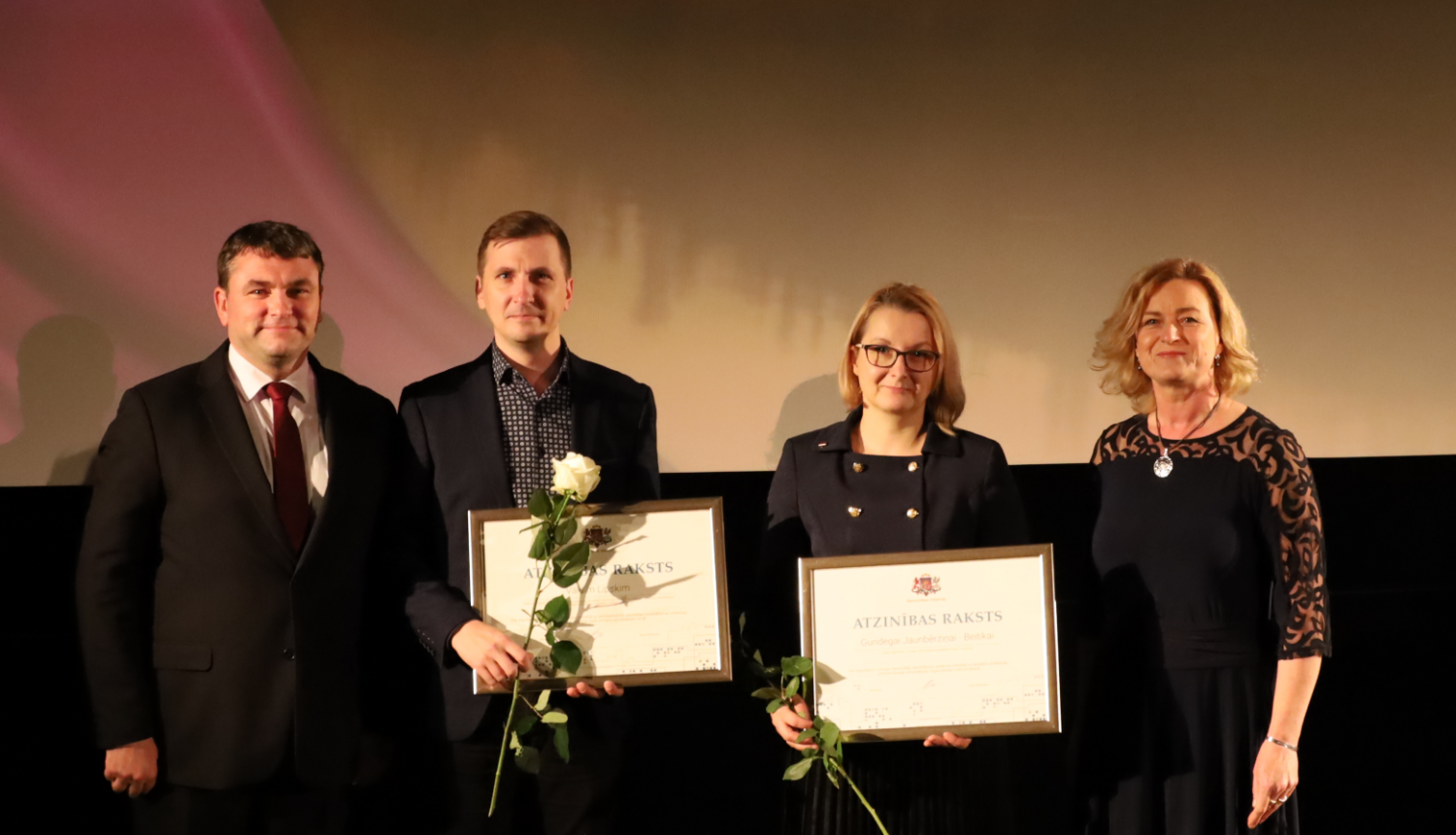 Četri cilvēki, divi no viņiem tur Atzinības rakstus. Divas sievietes, divi vīrieši. Tumšs fons