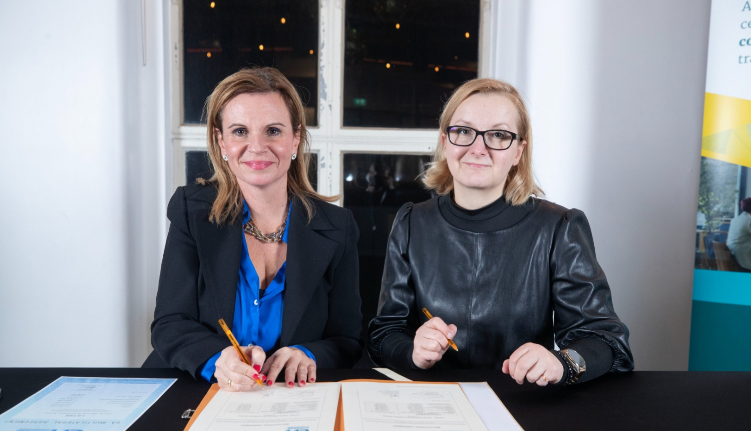 Parakstīšanas brīdis, kurā piedalās LATAK direktore un EA pārstāve. Uz galda ir mape ar dokumentiem un abas pārstāves veic parakstīšanu.